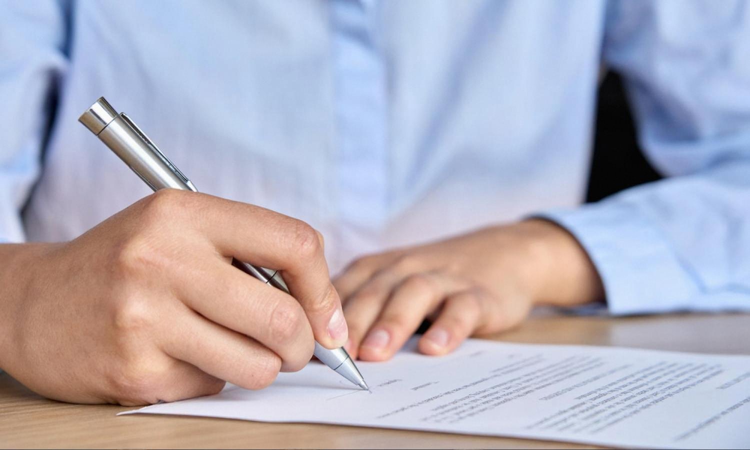 Close-up of a person in a light blue shirt signing a company contract with a silver pen